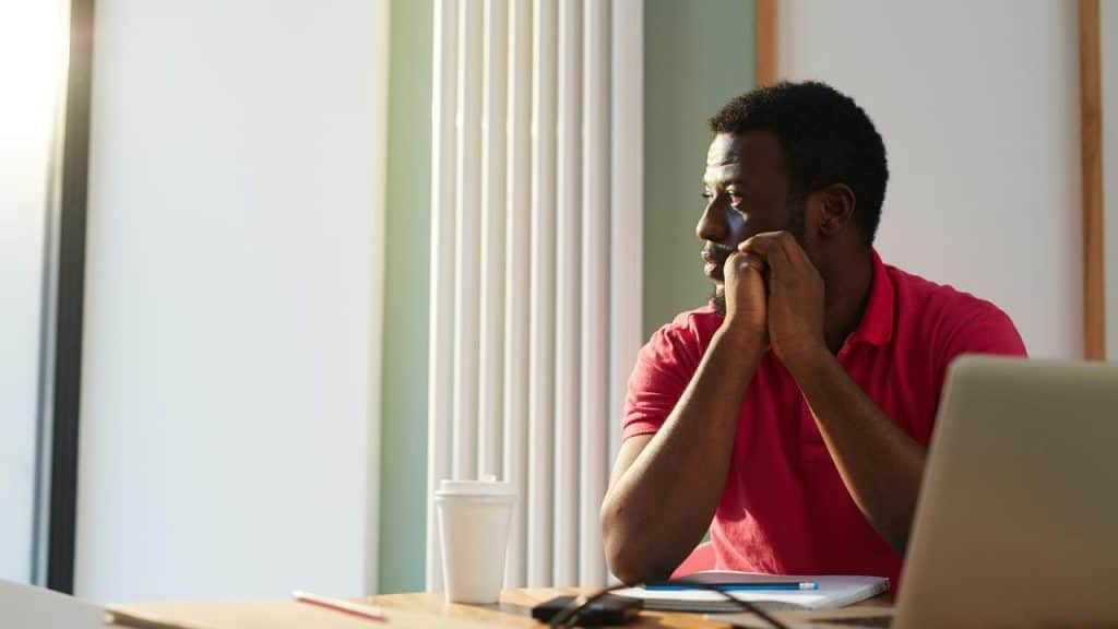 A man in a red shirt sitting at a desk, looking thoughtfully out the window.