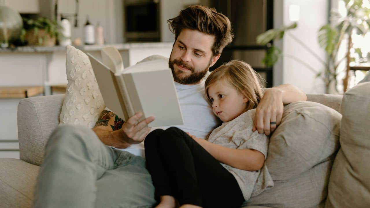 Smiling man with a beard reading a book to a young girl on a sofa.