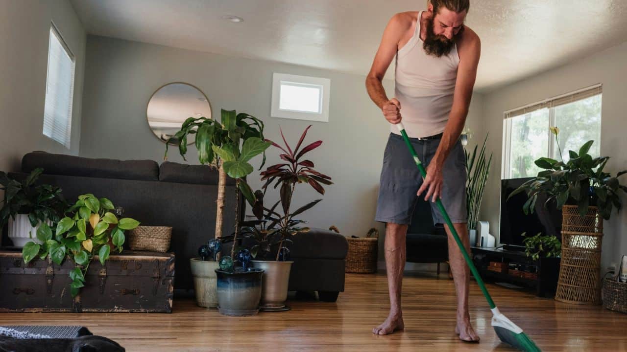 Bearded man in tank top and shorts sweeping a wooden floor in a sunlit living room.