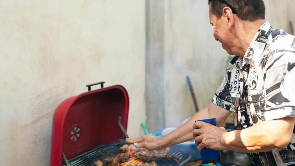 A man grills meat on a red barbecue while holding a drink and smiling outdoors.