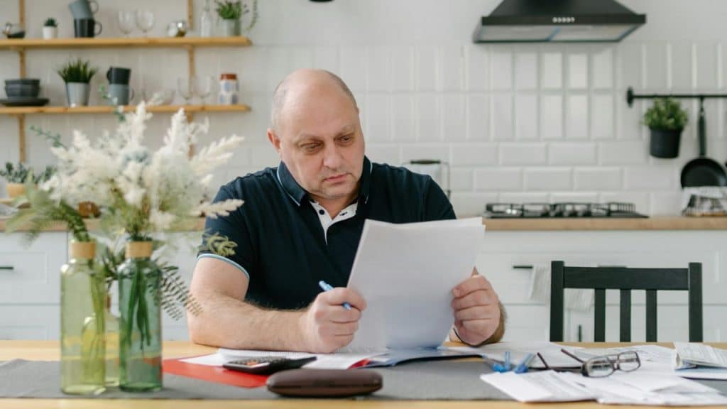 Bald man sitting at a kitchen table looking seriously at a paper document.