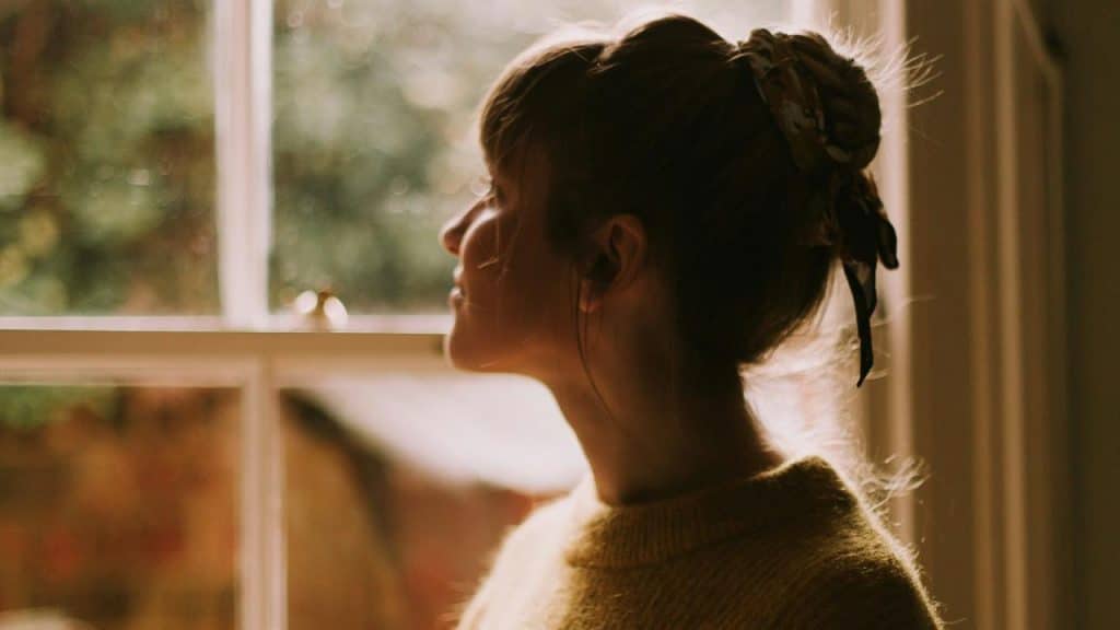 A woman with her hair tied up looks out of a window thoughtfully.