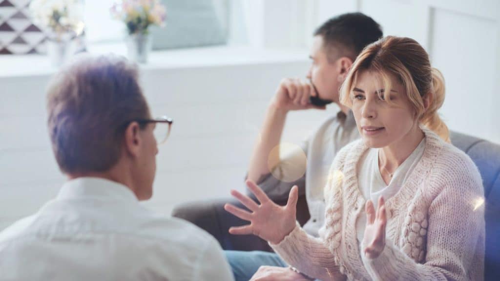 Woman gesturing while speaking to a man in glasses; another man is seated in the background.