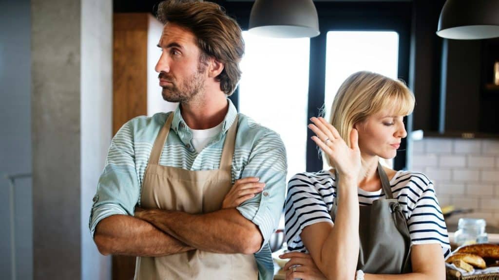 A man and woman, both wearing aprons, stand back-to-back in a kitchen.