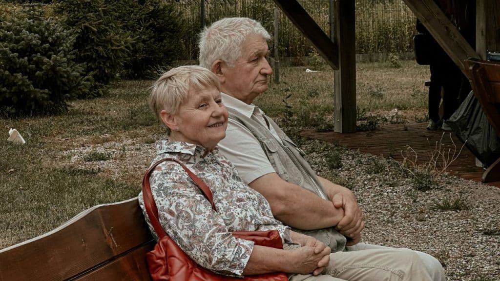 Elderly couple sitting side-by-side on a bench outdoors, woman smiling.