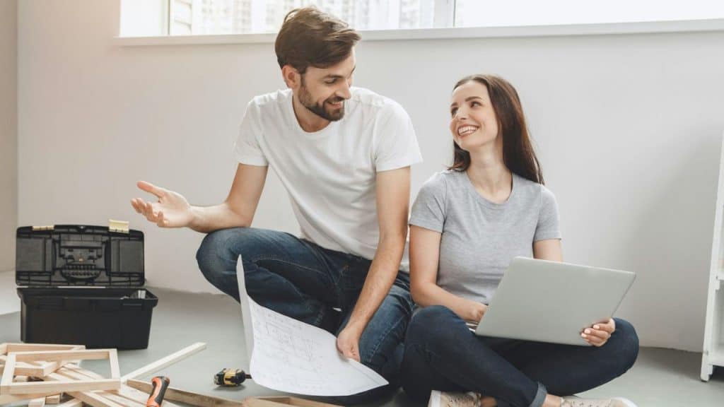 Smiling couple sitting on the floor with a laptop and blueprints, surrounded by tools.