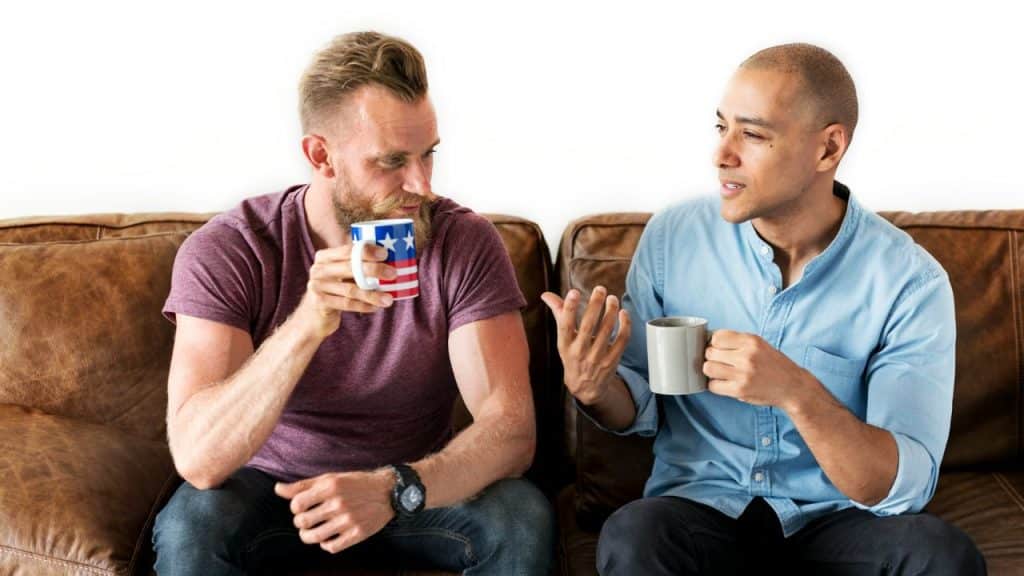 Two men sitting on a brown couch talking, each holding a coffee mug.