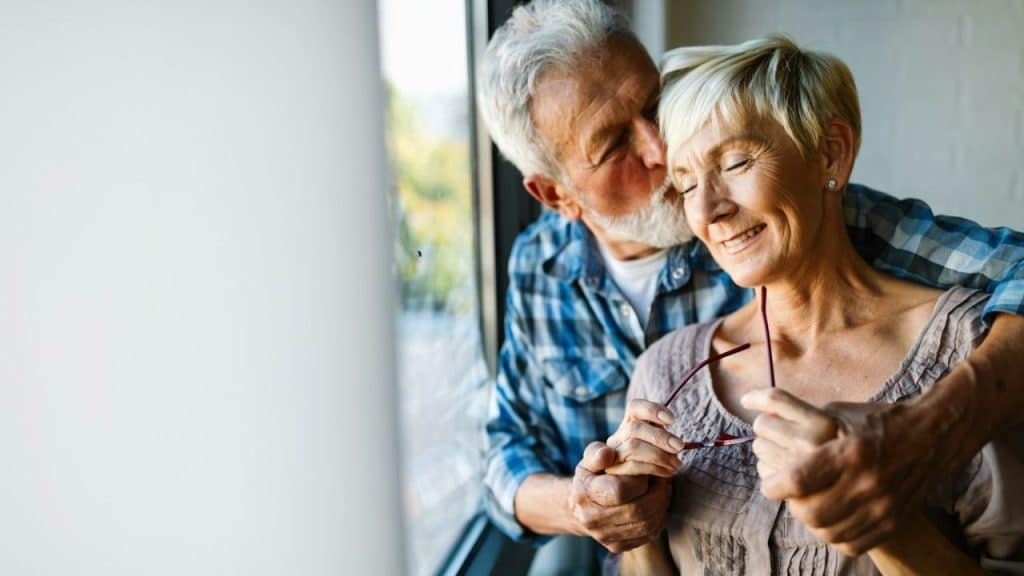 An older man kisses a smiling woman's cheek as they stand near a large window.