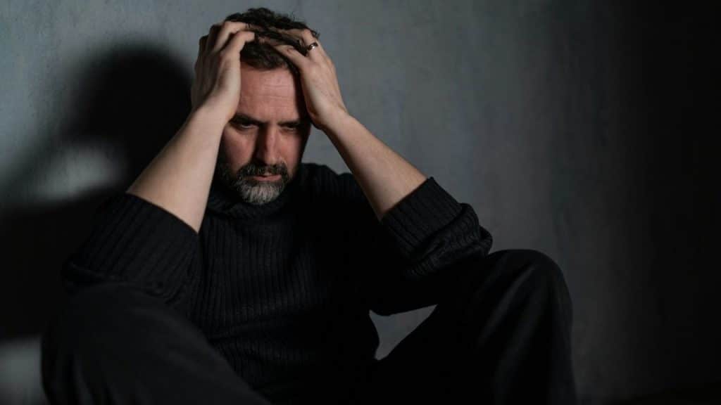 Middle-aged man in a dark room holding his head in his hands, looking distressed.
