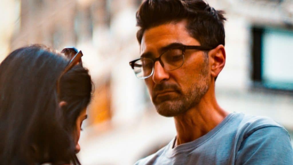 Man with glasses and a beard looking down sadly while talking to a woman.