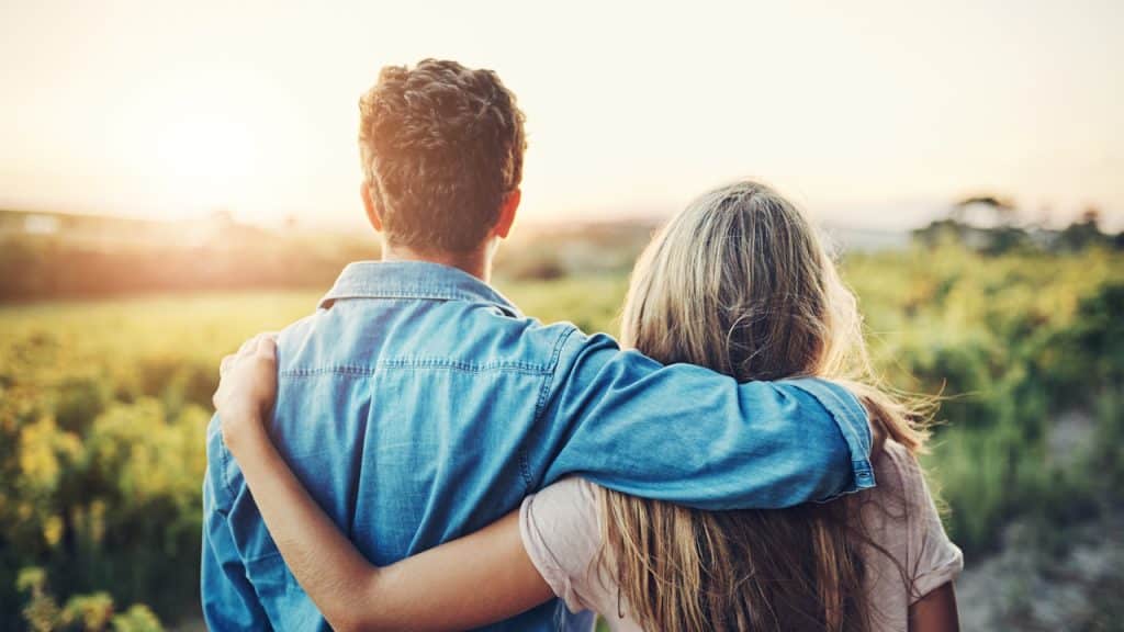Couple with arms around each other looking out over a sunny field.