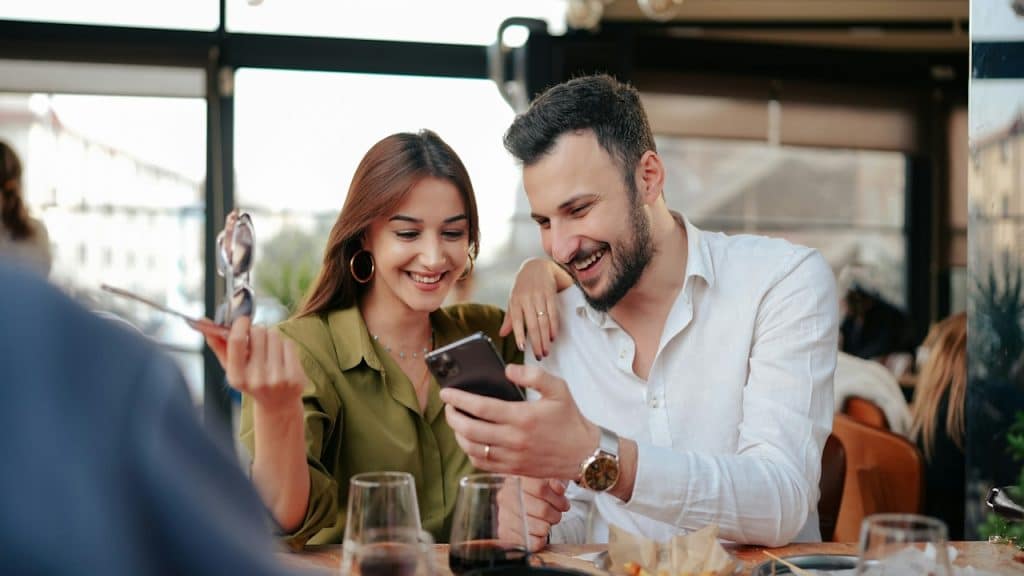A man showing a woman holding sunglasses something on his phone while they sit at a dinner table. 