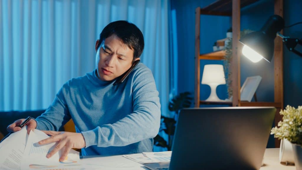 A man working on some papers with his laptop placed before him on a table.