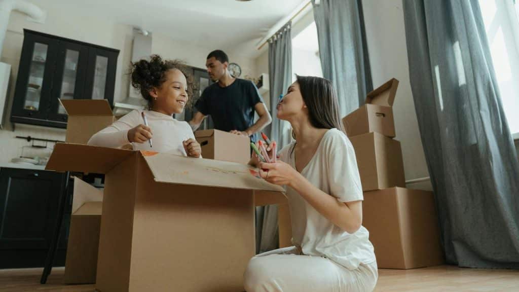 A woman and a girl play with markers in a moving box while a man unpacks in the background.