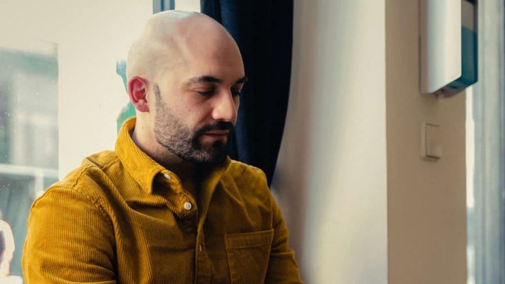 A bald man with a beard, wearing a mustard yellow corduroy shirt, looks down.