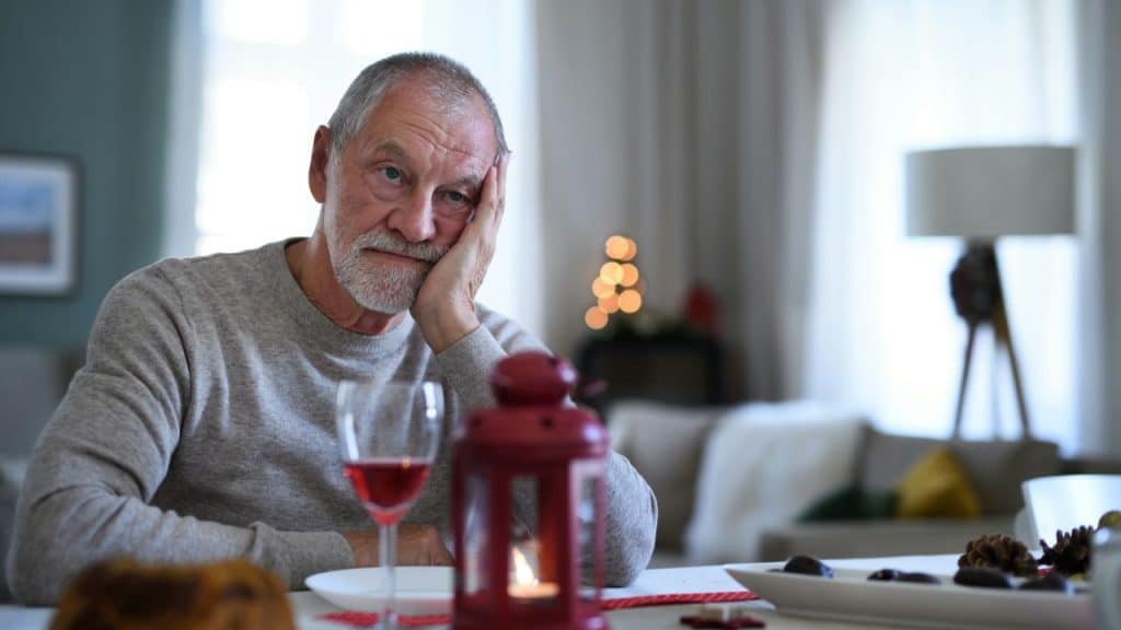An older man looks sadly at the camera while sitting at a table with a glass of wine.
