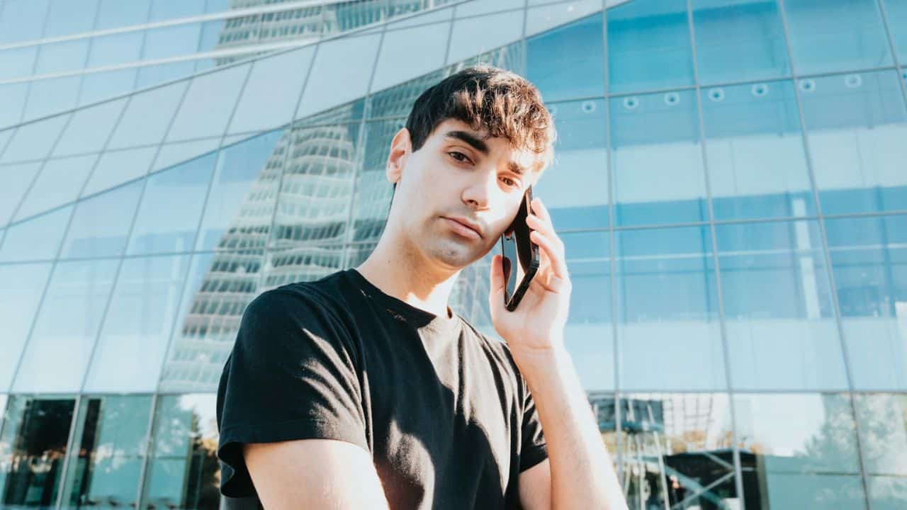 A young man on a cell phone stands outside a modern glass skyscraper on a sunny day.