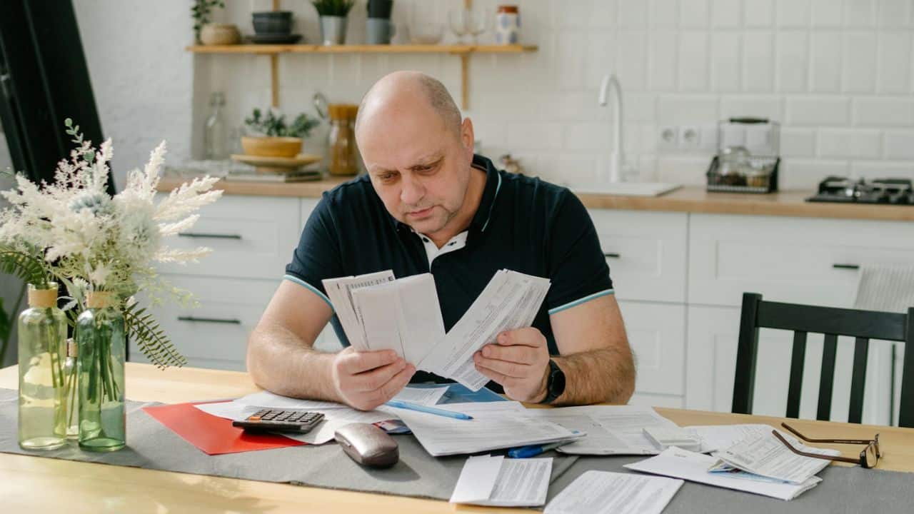Bald middle-aged man intently reviewing multiple paper documents at a kitchen table.
