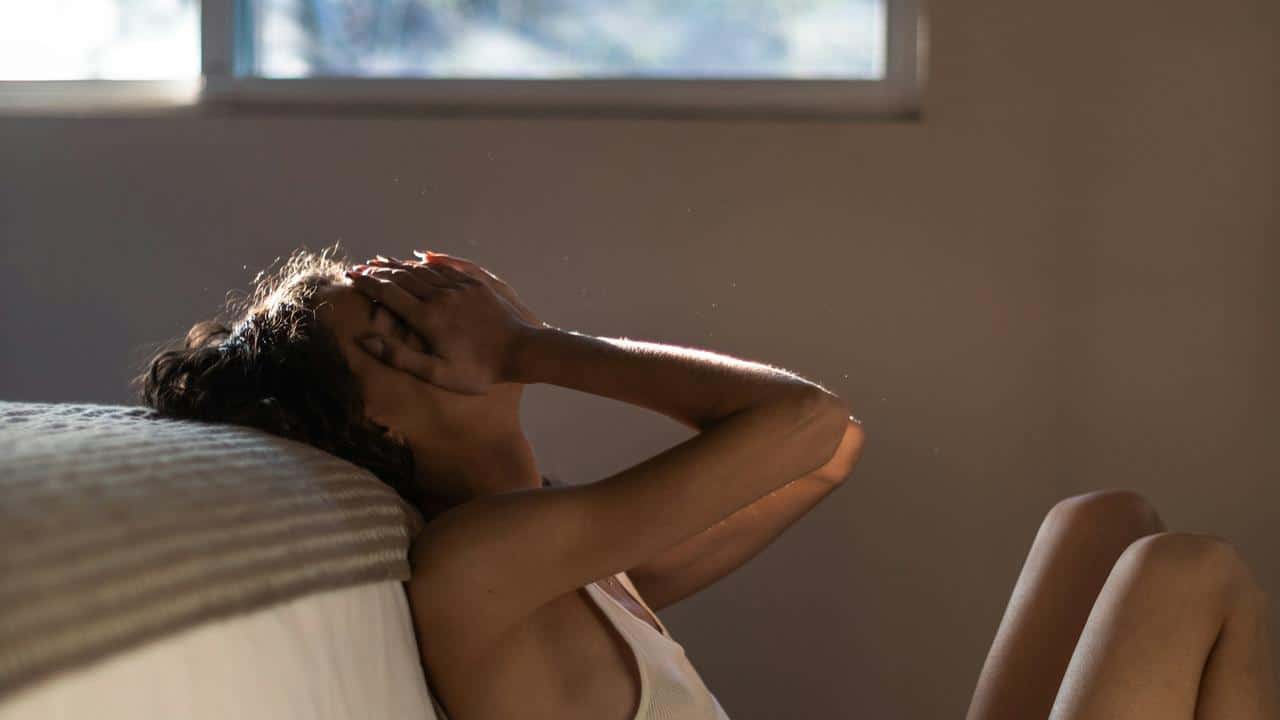 A woman sitting on the floor by a bed with her hands covering her face.