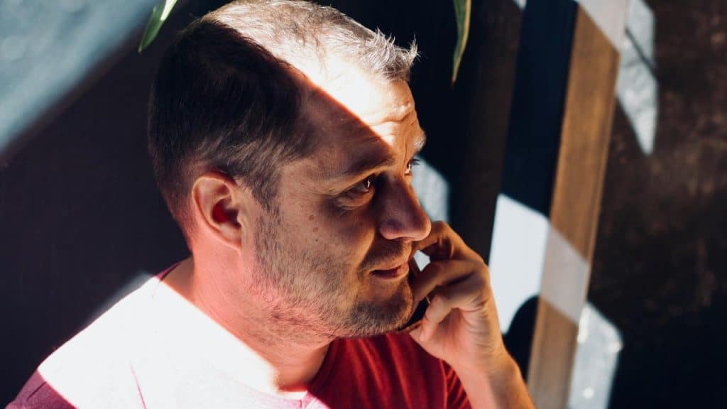 A man in a red shirt talks on the phone while sunlight falls across his face.