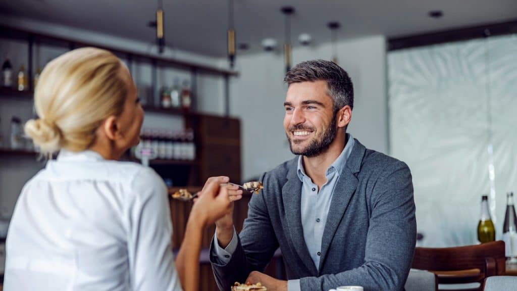 Smiling man being fed a bite of dessert by a woman across a cafe table.