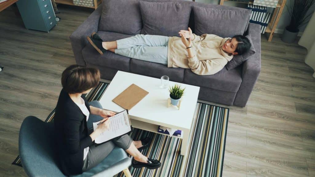 A person lying on a couch talking with a therapist taking notes.