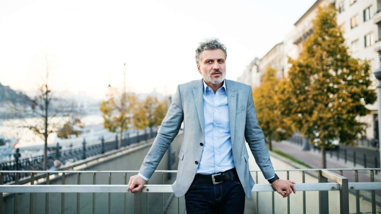 A man in a blazer is standing and leaning on a railing in a city during autumn.