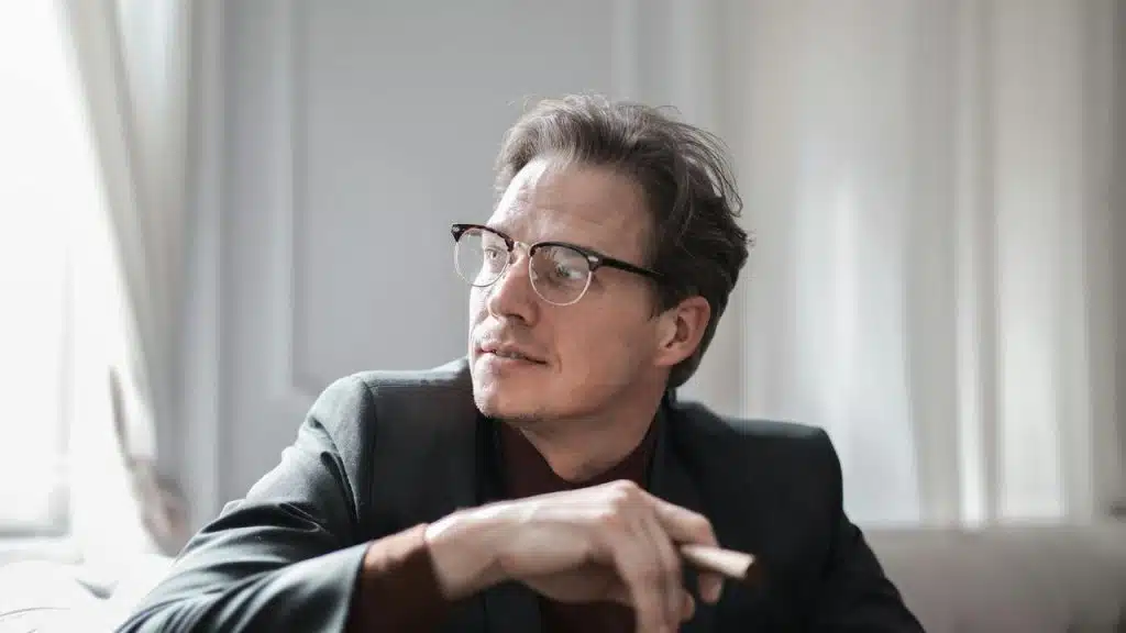 A man in glasses looking to the side while holding a cigar in a softly lit room.