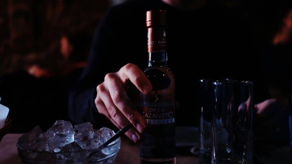 A hand reaching for a bottle of Ouzo on a dark table, next to a bowl of ice and empty glasses.