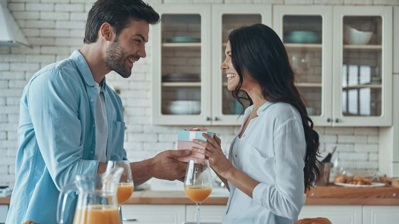 A smiling man is handing a small gift box to a woman in a bright kitchen.