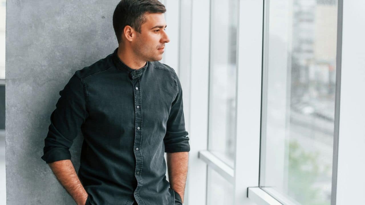 A man in a black collared shirt is standing by a large window, looking to the side.
