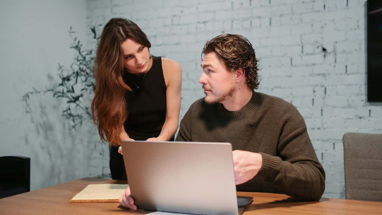 A woman leans over to talk to a man sitting at a desk with a laptop.