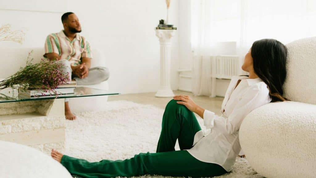 A woman sits on a shag rug, looking back at a man seated on a white sofa.
