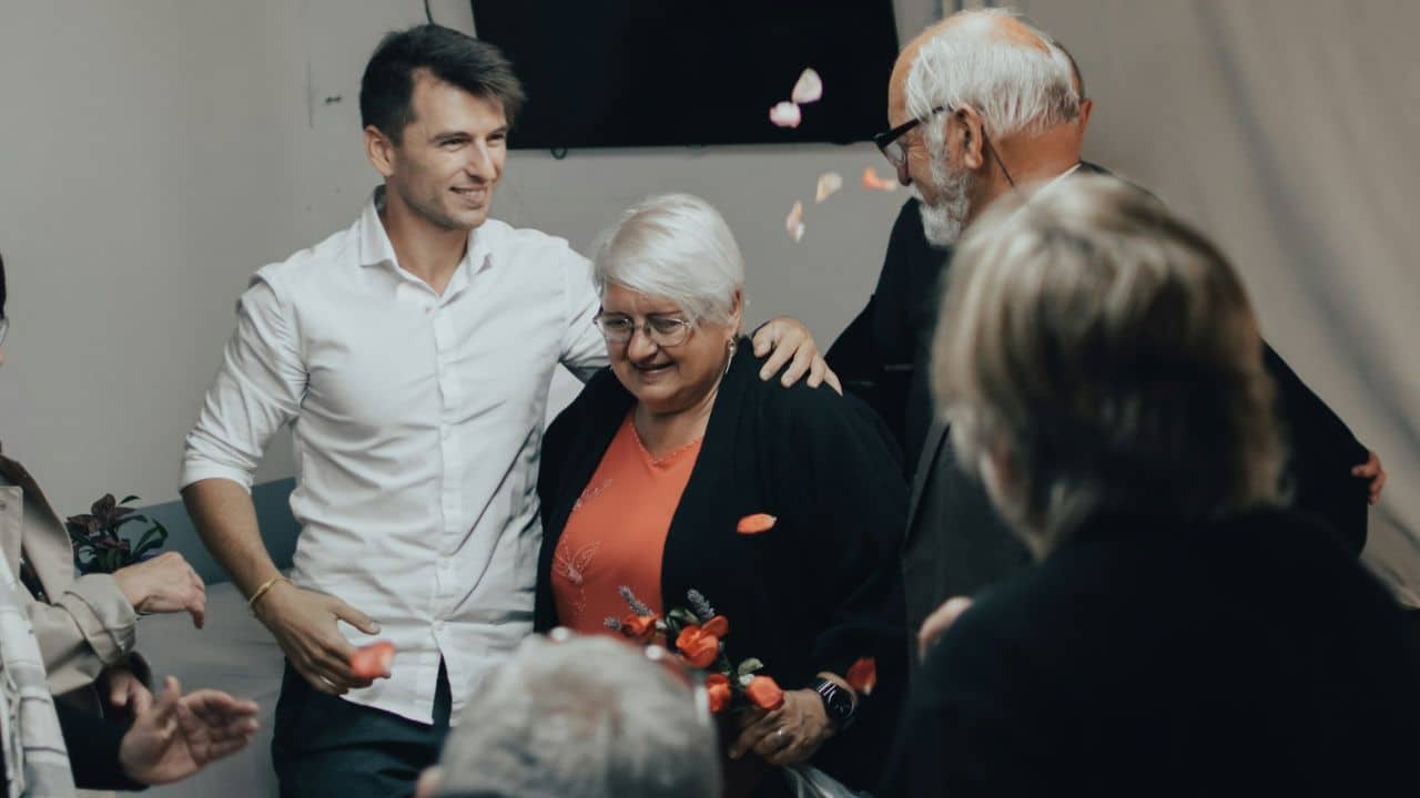 A group of people, including an older woman with glasses, are smiling indoors.