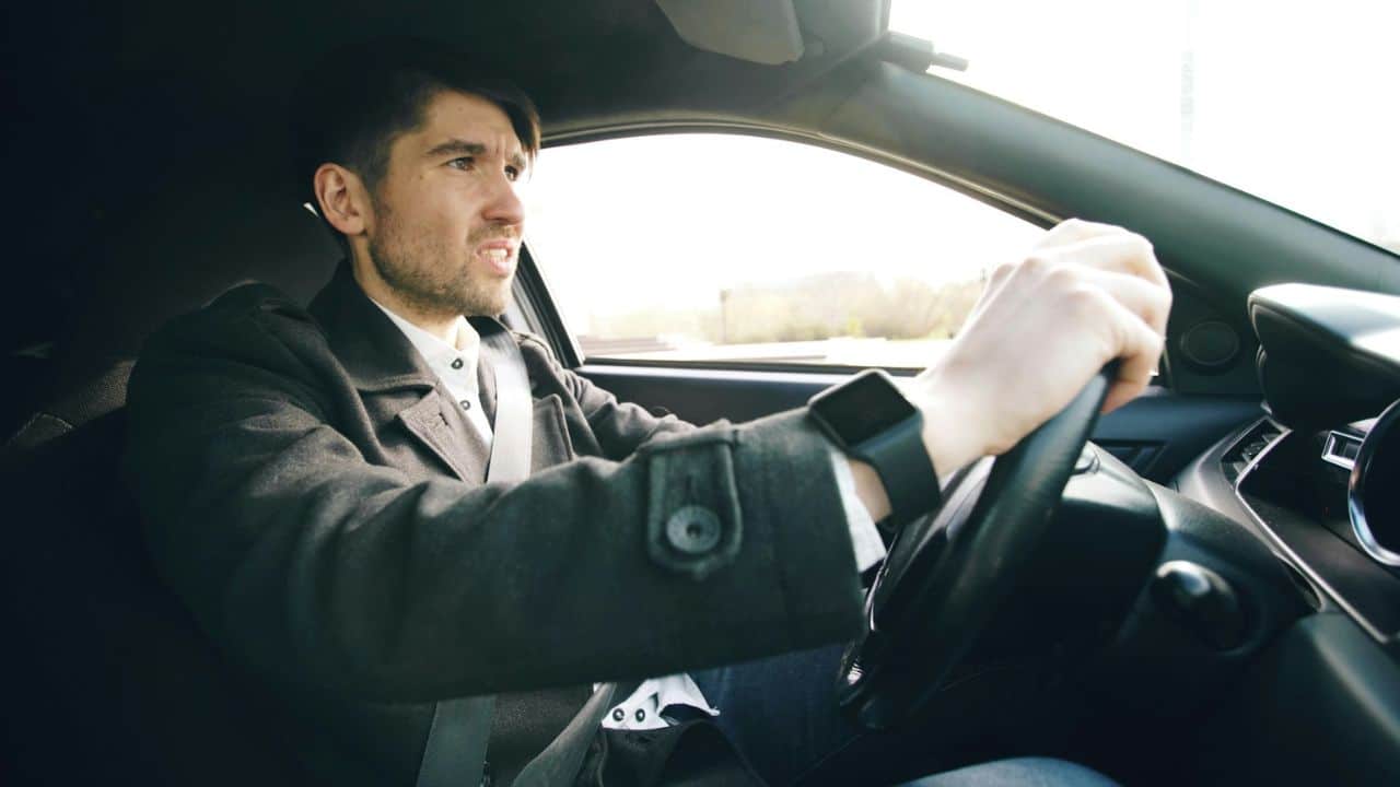 A man in a coat and seatbelt drives a car, gripping the steering wheel.