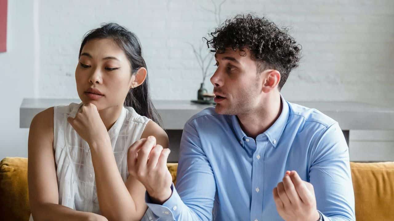 A man with curly hair gestures while talking to a woman sitting next to him.