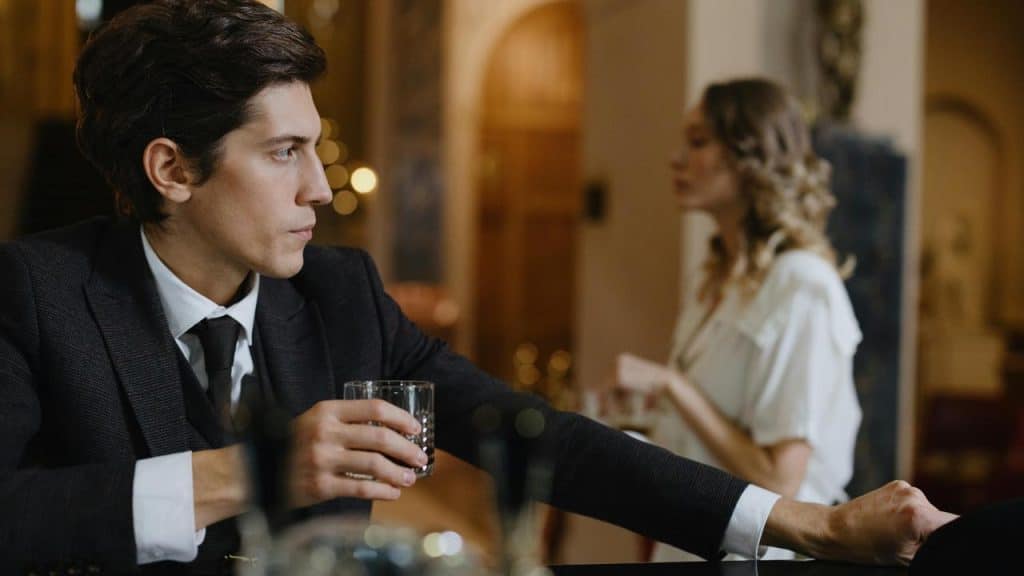 A man in a suit holding a drink at a bar with a woman blurred in the background.