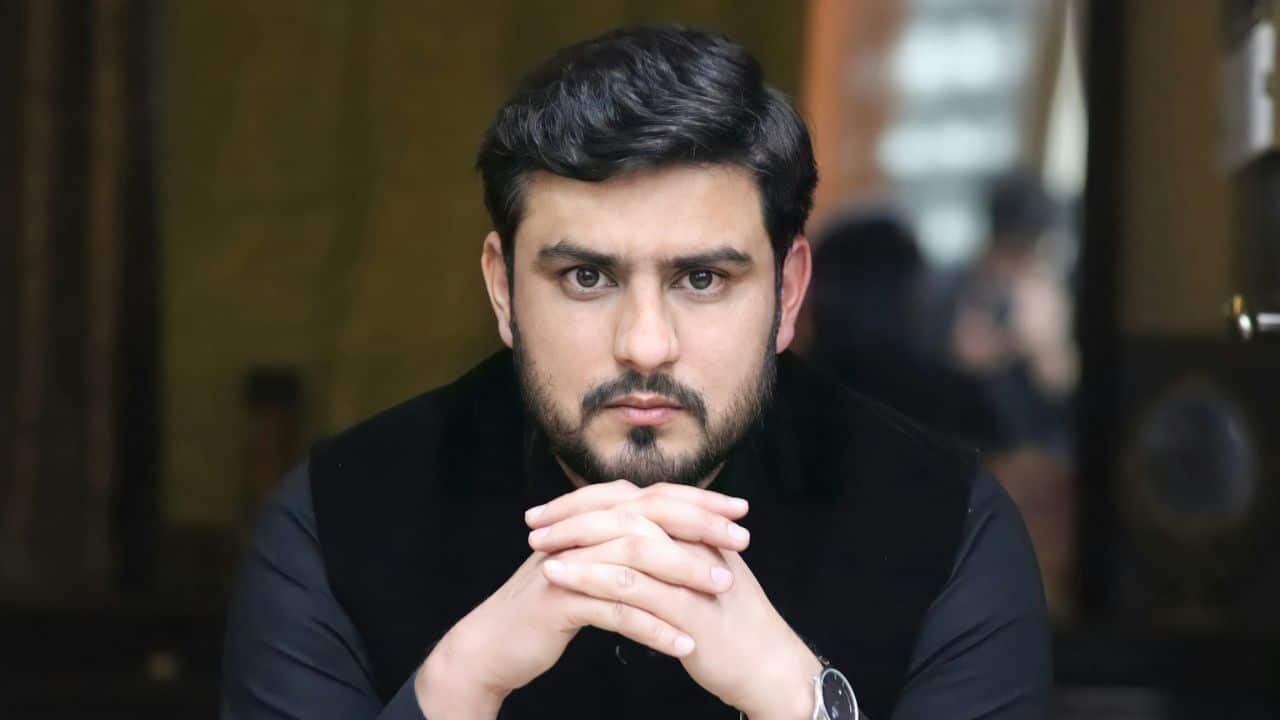 A bearded man in a black vest and shirt stares directly at the camera with his hands clasped.