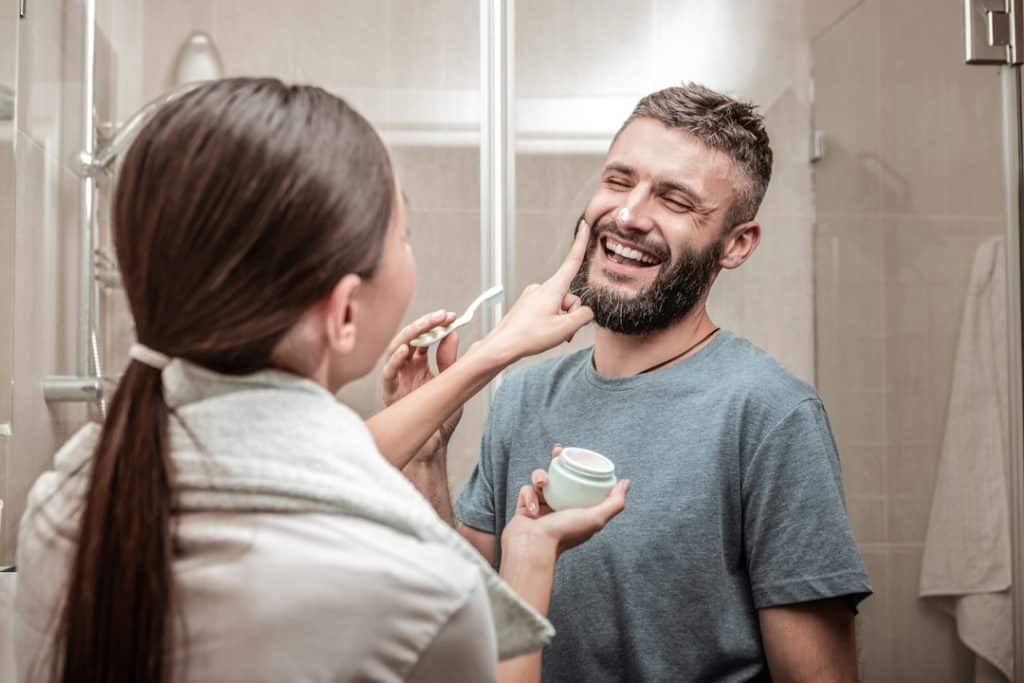 A caring girlfriend standing in front of her boyfriend while putting cream on his face.