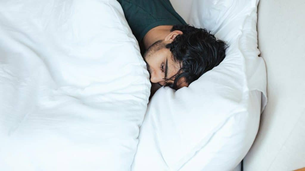 A man lying in bed with his head on a pillow, partially covered by a blanket.