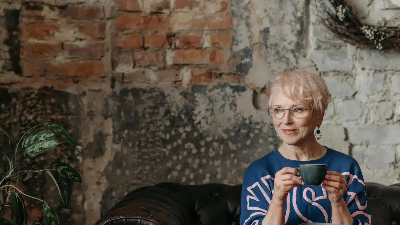 An older woman sitting against a rustic wall while holding a cup.