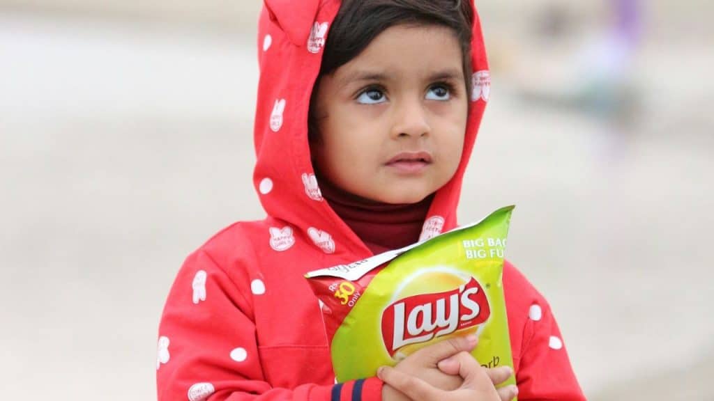 A child in a red hooded jacket holds a bag of Lay's chips, looking up.