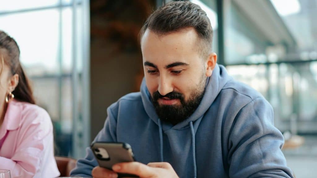 A bearded man in a blue hoodie looks down at his phone in an outdoor setting.