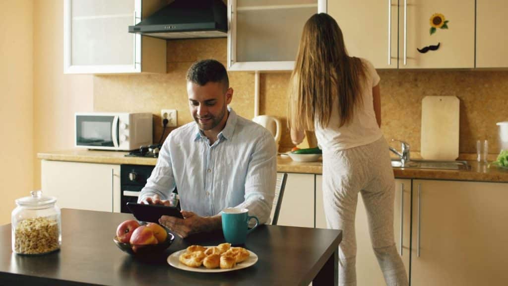 Man sits at a kitchen table using a tablet while a woman stands, facing away, washing dishes.