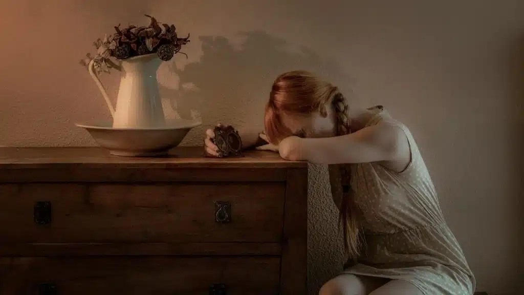 Woman with red braids leaning her head on a wooden dresser in dim light.