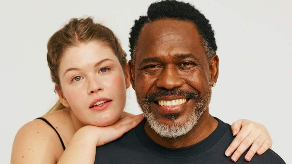 An older bearded man smiling while a younger woman stands at his back with her arms wrapped around his shoulders. 