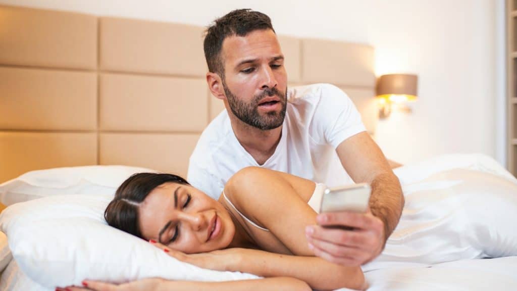 A man in bed is looking intensely at a smartphone he is holding, while a woman sleeps next to him.