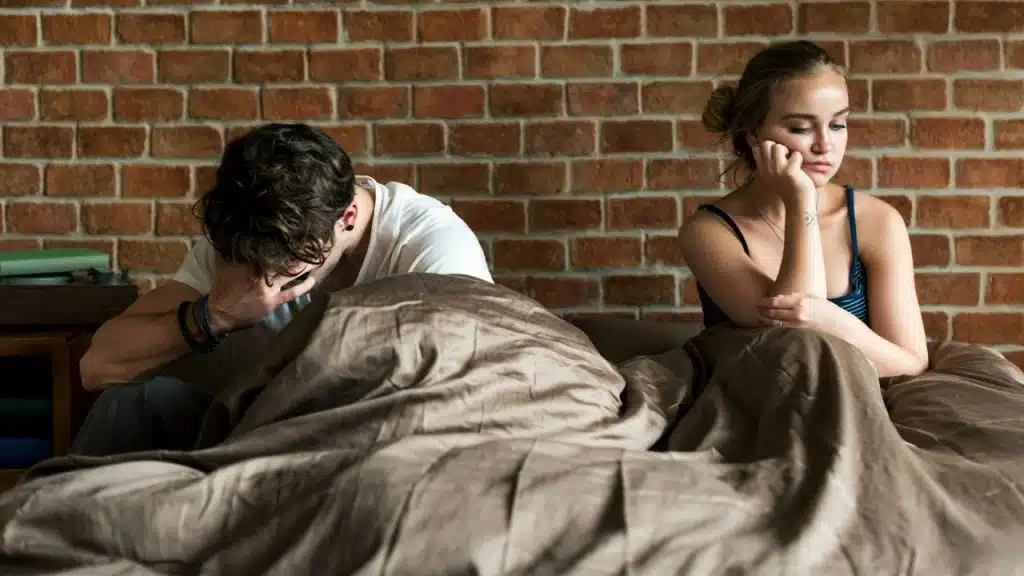 Man and woman sitting apart on a bed with brown bedding and brick wall.