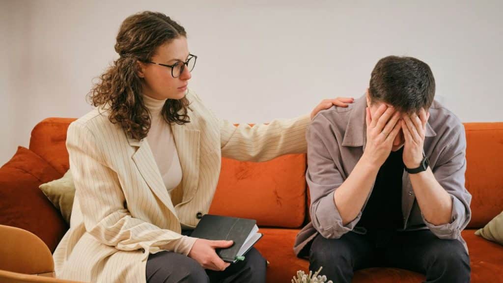 Woman comforting a distraught man covering his face while sitting on an orange couch.