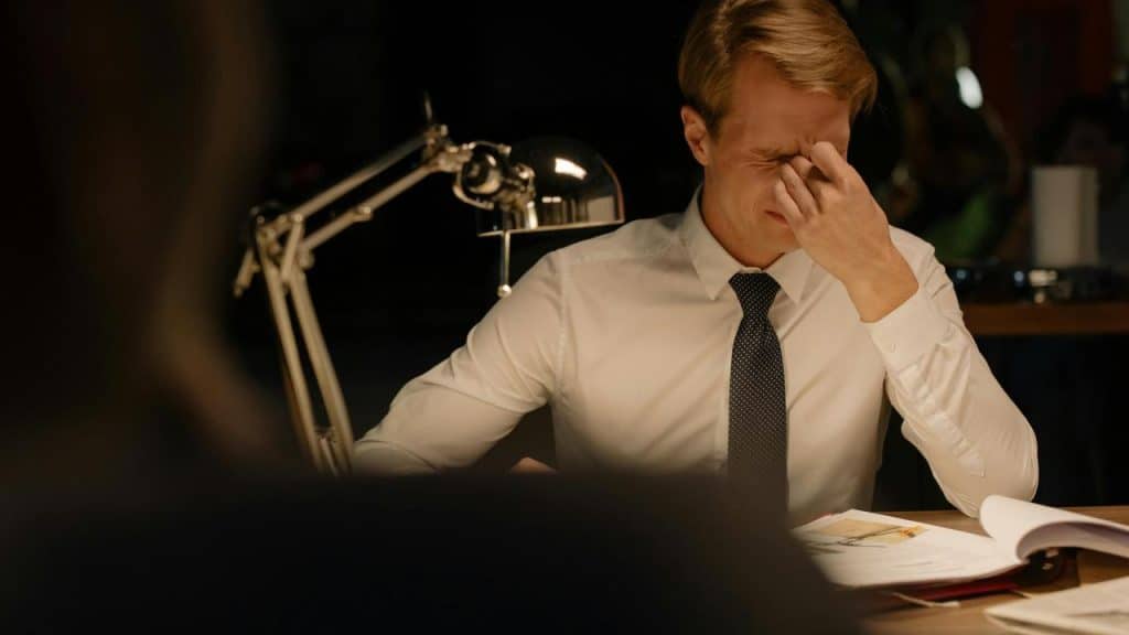 A man in a white shirt and tie sitting at a desk, pinching his nose in frustration.